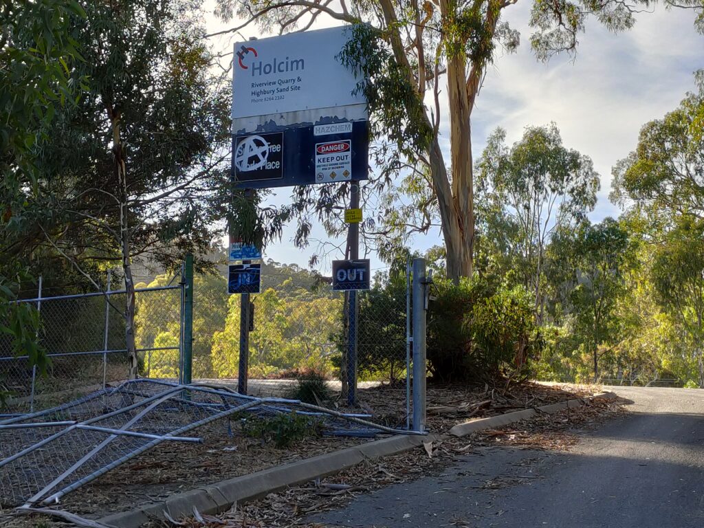 Photograph of trees and fallen-down cyclone fence in front of a sign marked 'Holcim-Riverview Quarry & Highbury Sand Site, HAZCHEM-DANGER KEEP OUT' covered in graffiti.