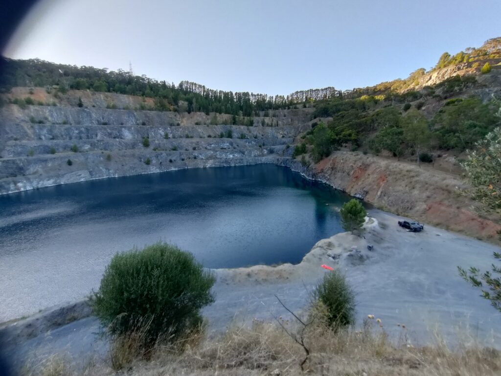 Photograph of large rocky quarry with trees on the upper slopes, filled with water and a car parked near the water's edge.