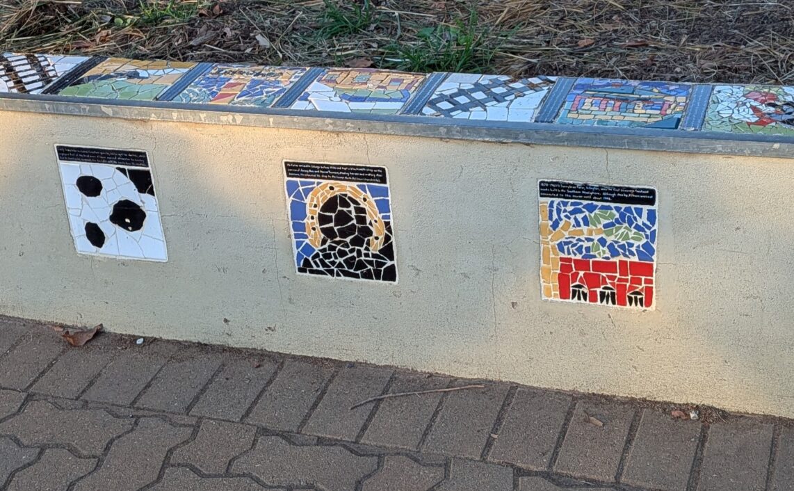 A concrete wall with mosaic tiles on top and sides showing iconic scenes from the site of Kilburn.