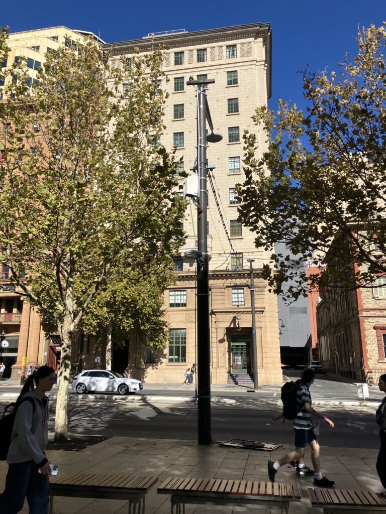 People walking past the camera in the shade, with benches in the foreground and colonial buildings in the background.