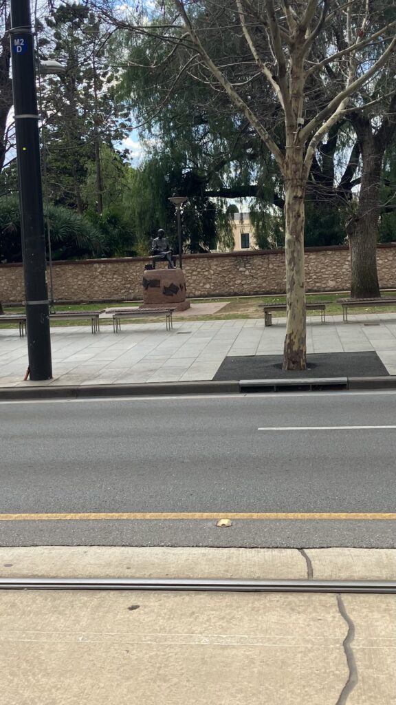Empty street with benches in the foreground and a statue of woman seated on a bench writing on her lap against a stone wall in the background.