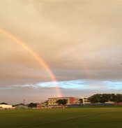 A rainbow appears in the sky above some large buildings and an oval.